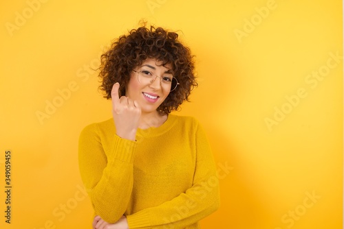 Young Arab woman with curly hair wearing yellow sweater  standing over isolated yellow background Beckoning come here gesture with hand inviting welcoming happy and smiling