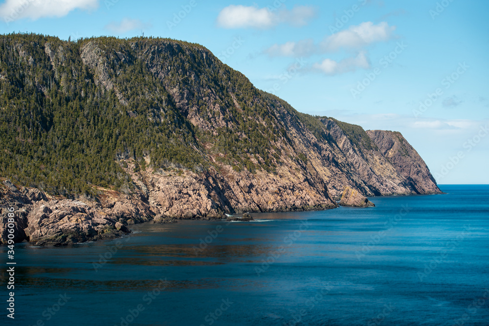 A large rocky mountain of green trees and rocky cliffs. The hill has a ragged and rough coastline. The ocean is blue and smooth below the mountain. The background is blue sky and white fluffy clouds.