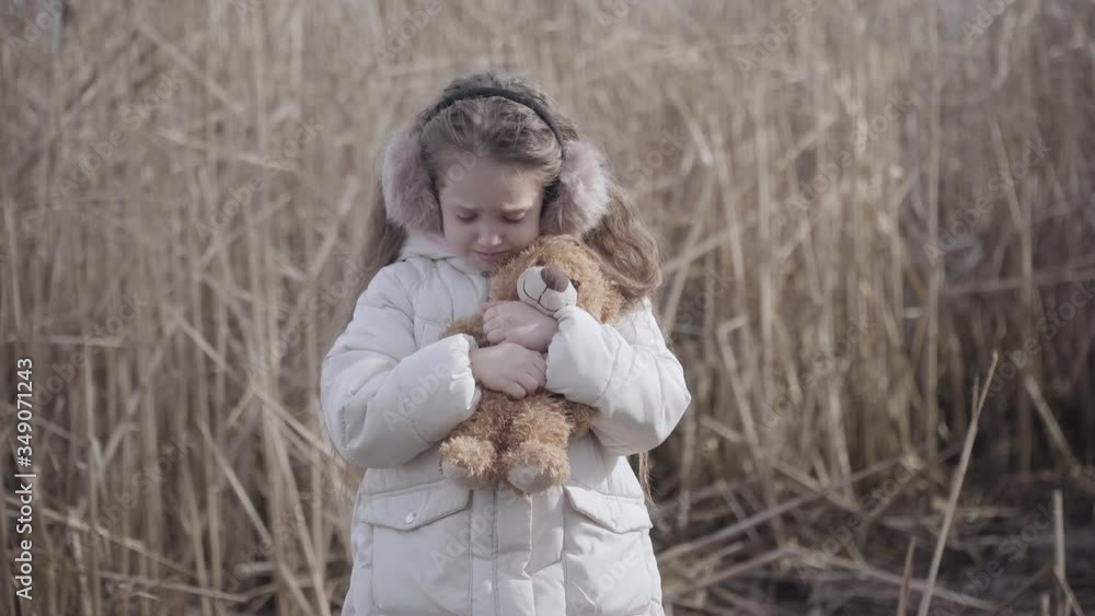 Portrait of lost Caucasian little girl with teddy bear crying outdoors. Sad lonely child standing at the background of grey bushes and hugging toy. Problem, depression, risk, abandonment. Portrait of lost Caucasian little girl with teddy bear crying outdoors. Sad lonely child standing at the background of grey bushes and hugging toy. Problem, depression, risk, abandonment.