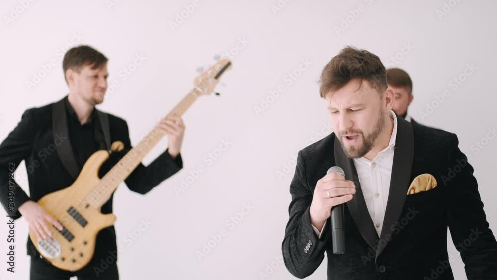 Men wear black suits. Vocalist holds microphone. Two men play musical instruments. Men wear black suits. Vocalist holds microphone. Two men play musical instruments.