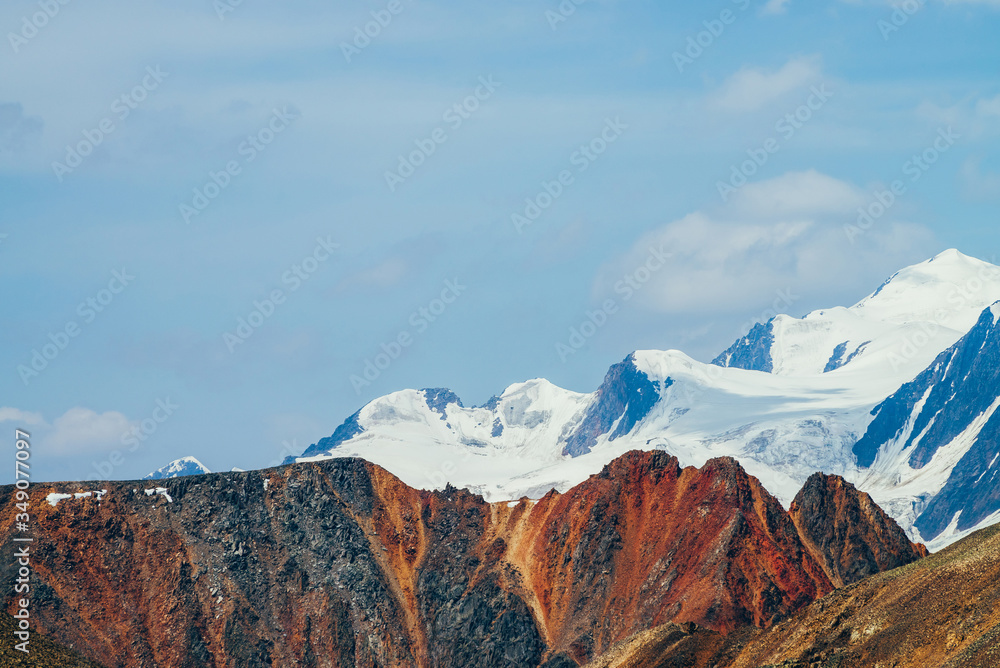 Minimalist beautiful alpine landscape with huge snowy mountain behind vivid pointed red craggy wall. Big glacier under blue sky. Atmospheric colorful highland scenery with giant red orange brown rock.