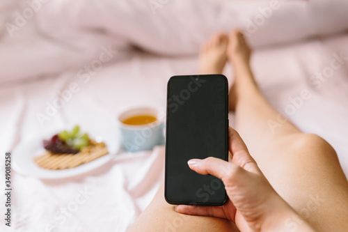 Young woman having a breakfast at home in her bed, checking her mobile phone. Focus on cell phone