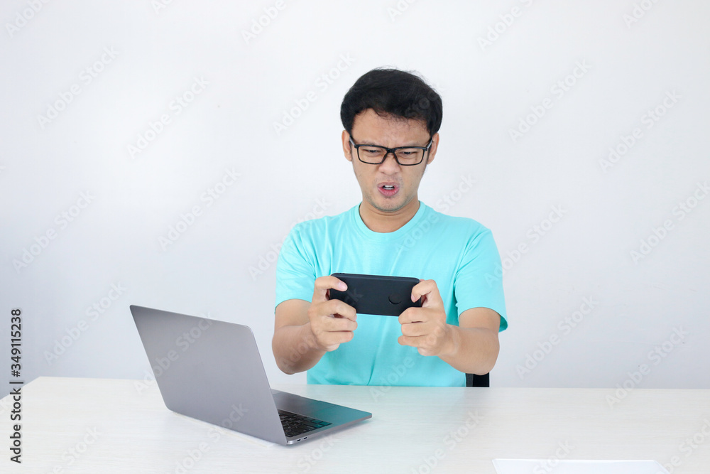 Wow face of Young Asian man shocked what he see in phone with laptop beside it. Indonesian man wearing blue shirt.