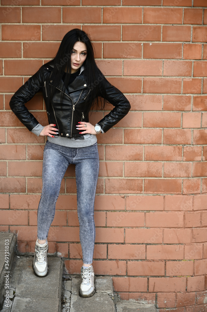 Full-length portrait in spring weather outdoors, caucasian brunette girl in jeans and a black jacket with a great figure. Model posing against the backdrop of a building wall. Full-length portrait in spring weather outdoors, caucasian brunette girl in jeans and a black jacket with a great figure. Model posing against the backdrop of a building wall.