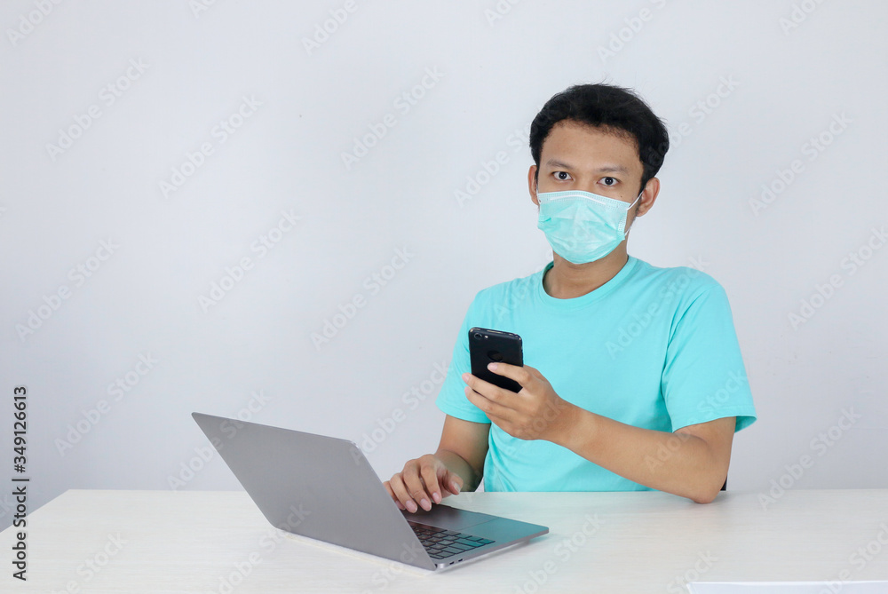 Young Asian Man wearing medical mask is serious and focus when working on a phone and laptop on the table. Indonesian man wearing blue shirt.