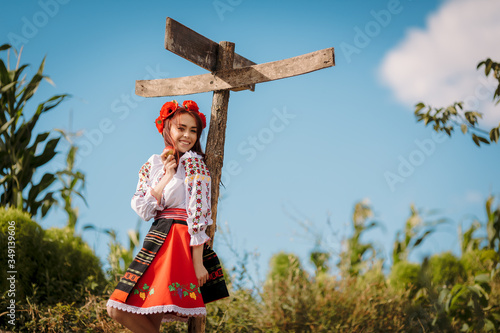 Young redhead moldavian girl dressed in traditional costume  with flower wreath on the head  staing near old pole with empty derections.