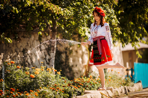 Young redhead moldavian girl dressed in traditional costume with flower wreath on the head watering  flowers