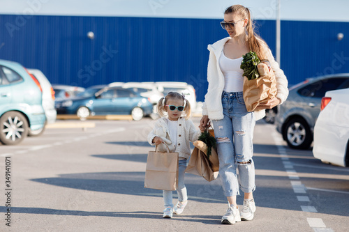 Mom and little daughter with ponytails, dressed in white T-shirts and blue jeans posing in the background of the parking lot near the supermarket with packages in hand. Mother and daughter shopping .