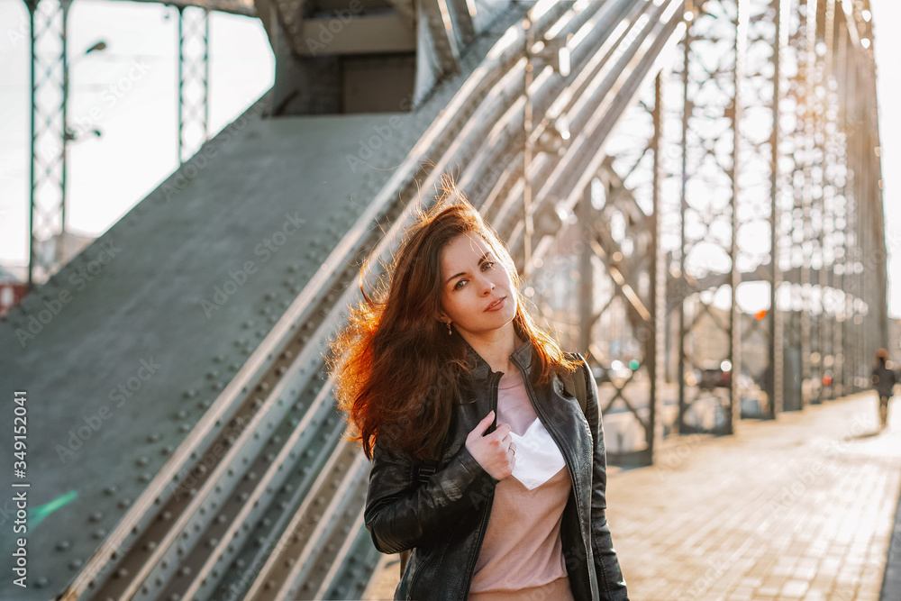 A young girl with hair flying in the wind in a leather jacket and a backpack walks on a metal bridge over the Neva river in St. Petersburg against the background of a highway in a Sunny beautiful suns