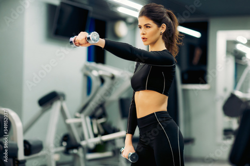 Young brunette girl, in black training leggings and long sleeve top goes in for sports with silver dumbbells in the gym. Free space for text. Concept of fitness club