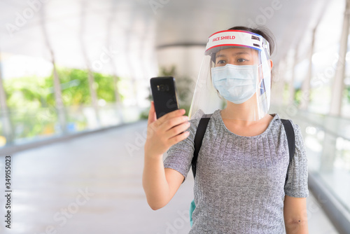 Young Asian woman using phone with mask and face shield for protection from corona virus outbreak in the city © Ranta Images