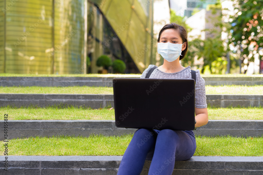 Young Asian woman with mask for protection from corona virus outbreak using laptop while sitting outdoors Young Asian woman with mask for protection from corona virus outbreak using laptop while sitting outdoors