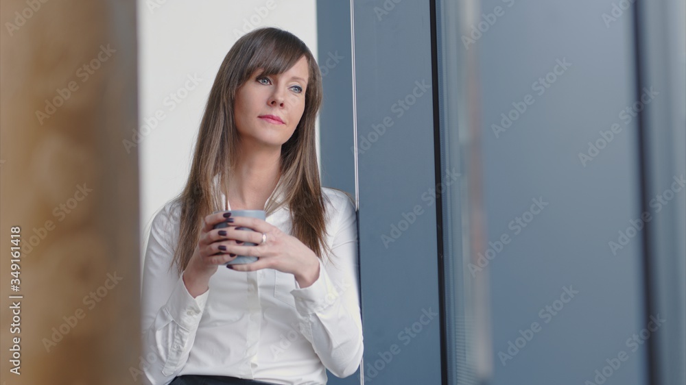 Businesswoman taking break in office drinking coffee standing in window and daydreaming, thinking.