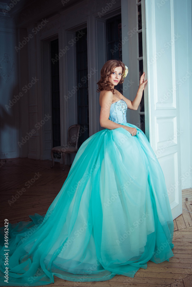 The beautiful young girl the princess in a long magnificent turquoise dress stands in a light castle. Fairytale fashion photo session Cinderella. Beautiful room. Princess runs at the door