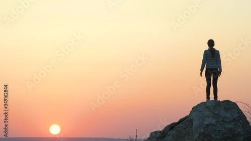 Silhouette of a woman hiker jumping alone on big stone at sunset in mountains. Female tourist raising her hands up on high rock in evening nature. Tourism, traveling and healthy lifestyle concept.
