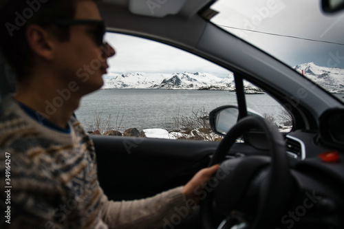 Man driving the car at the Lofoten islands, Norway. Cold fjord and snowy mountains view from window. Focus on the background © WellStock
