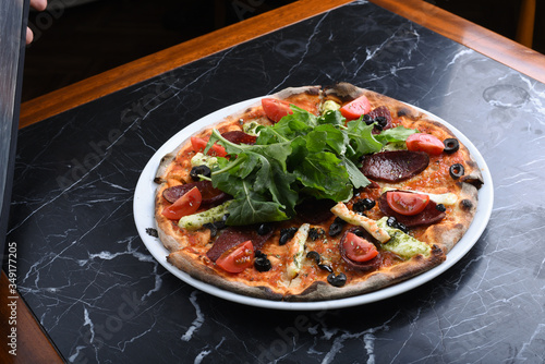 pizza with arugula decorated with sausage, black olive, tomato and cheddar cheese slices on a black marble table