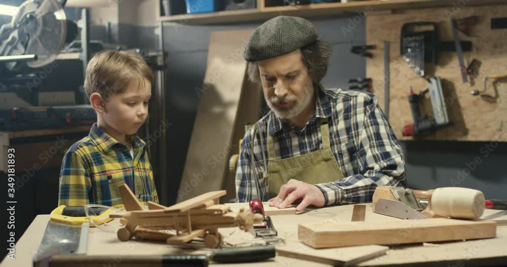 Caucasian woodman with gray beard teaching his little grandson to work with hardwood and saw timber. Old man carpenter in apron showing to small boy proceeding wood in workshop and filing File working Caucasian woodman with gray beard teaching his little grandson to work with hardwood and saw timber. Old man carpenter in apron showing to small boy proceeding wood in workshop and filing File working