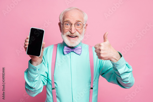 Portrait of amazed excited old man hold new smartphone show thumb up sign recommend suggest select wear teal turquoise shirt purple bowtie isolated over pink pastel color background
