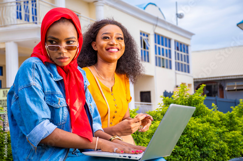 Quadro em tela pretty black girls sitting outdoor together using a laptop, one laughing and the