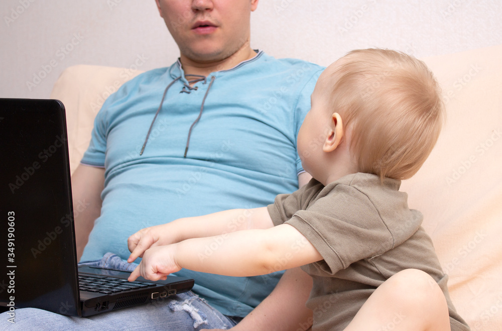 Naklejka premium Little baby sitting on father's knees at his workplace. Man holding child in front of laptop and working remotely.