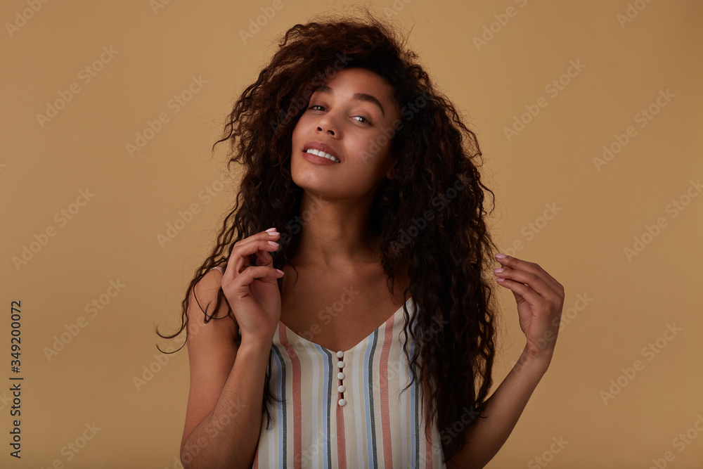 © timtimphoto - Cheerful young beautiful brown haired curly dark skinned lady with natural makeup looking positively at camera while standing over beige background with raised hands © timtimphoto - Cheerful young beautiful brown haired curly dark skinned lady with natural makeup looking positively at camera while standing over beige background with raised hands