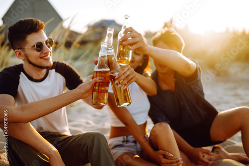 Group of young friends sitting together at the beach talking and drinking beers at sunset. Summer holidays, vacation, relax and lifestyle consept.