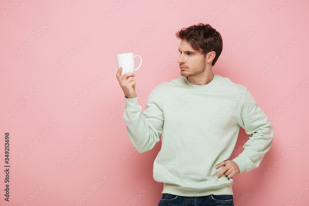 thoughtful young man looking at white cup while holding hand on hip on pink background thoughtful young man looking at white cup while holding hand on hip on pink background