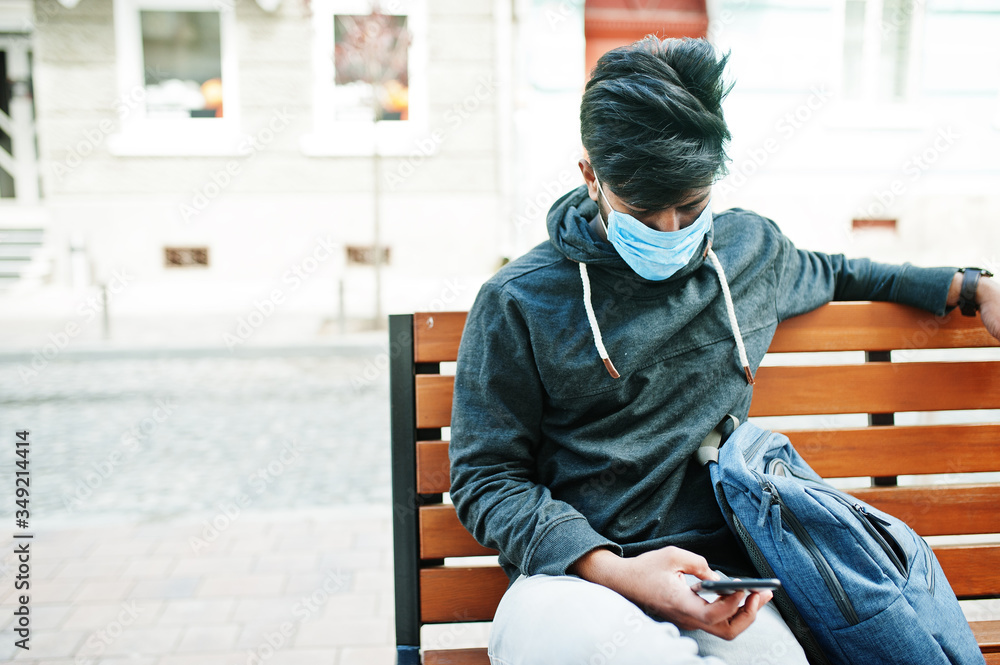 Coronavirus covid-19 concept. South asian indian man wearing mask for protect from corona virus sitting on bench with mobile phone.