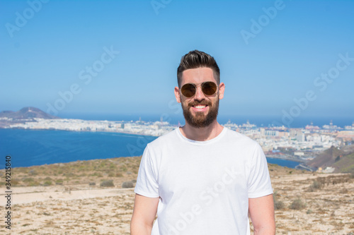 Handsome man with sunglasses  beard  and a white shirt on the top of the mountain from where you can see the edge of the island and the ocean  illuminated by sunlight.