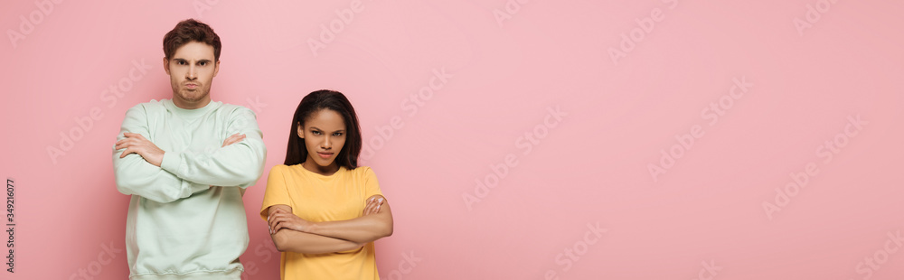 horizontal image of offended interracial couple standing with crossed arms while looking at camera on pink background horizontal image of offended interracial couple standing with crossed arms while looking at camera on pink background