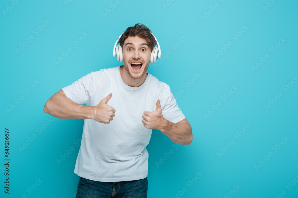 excited young man in wireless headphones showing thumbs up while looking at camera on blue background excited young man in wireless headphones showing thumbs up while looking at camera on blue background