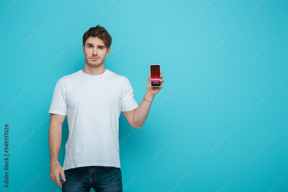 displeased young man showing smartphone with trading courses on screen on blue background displeased young man showing smartphone with trading courses on screen on blue background