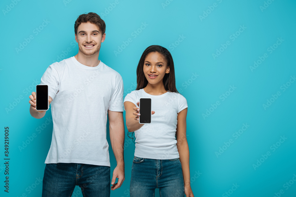 smiling interracial couple showing smartphones with blank screen on blue background smiling interracial couple showing smartphones with blank screen on blue background