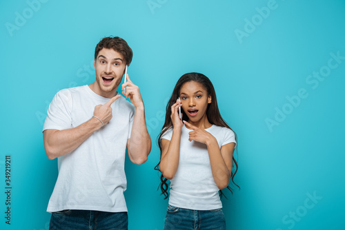 surprised interracial couple pointing with fingers while talking on smartphones on blue background