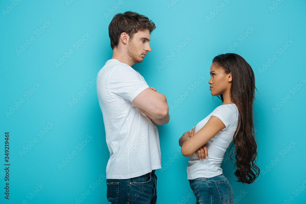 side view of offended interracial couple standing with crossed arms and looking at each other on blue background side view of offended interracial couple standing with crossed arms and looking at each other on blue background