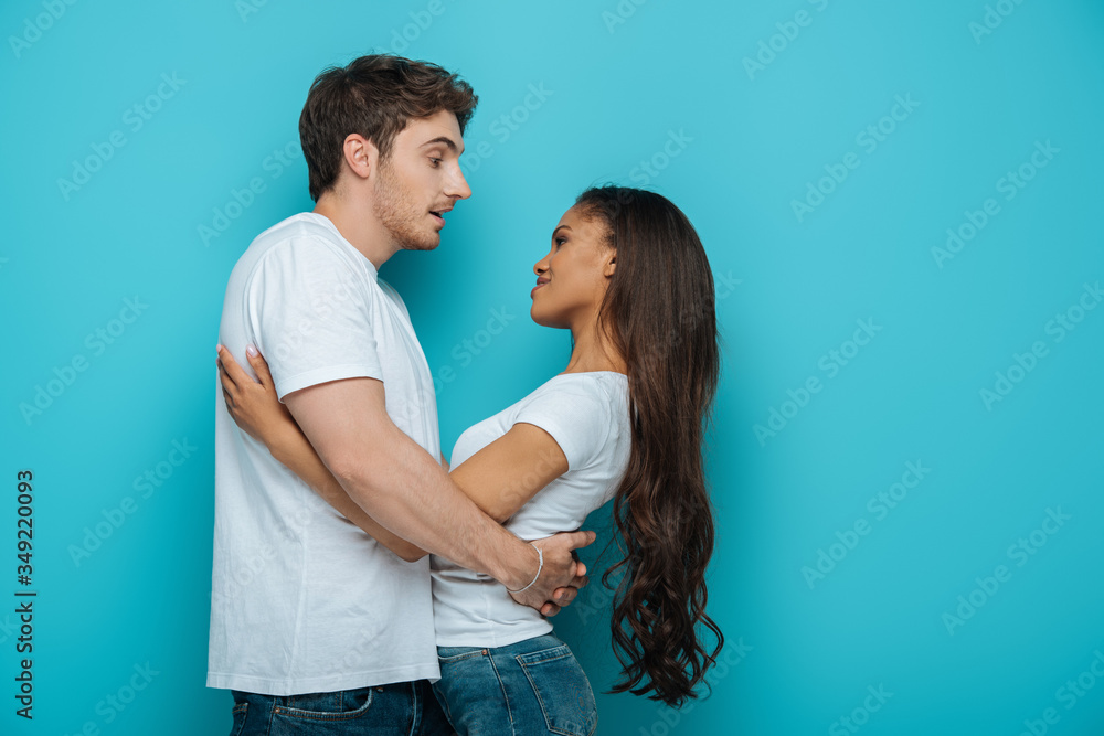 side view of young interracial couple hugging and looking at each other on blue background side view of young interracial couple hugging and looking at each other on blue background