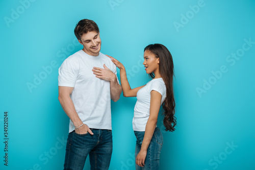 shy young man holding hand on chest while african american girl touching his shoulder on blue background