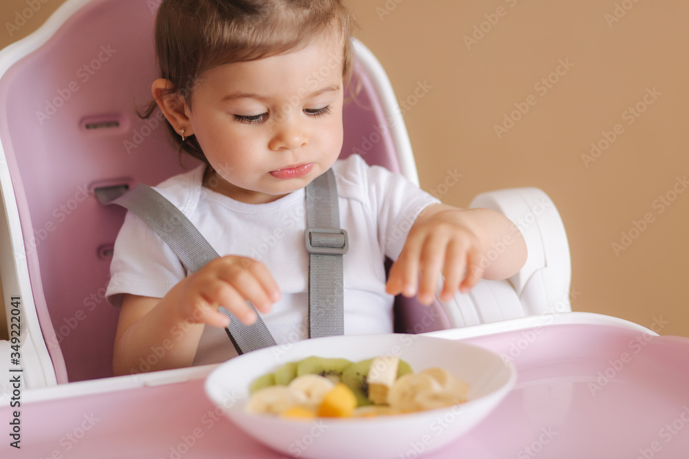 Portrait of happy young baby girl in high chair eating exotic fruits