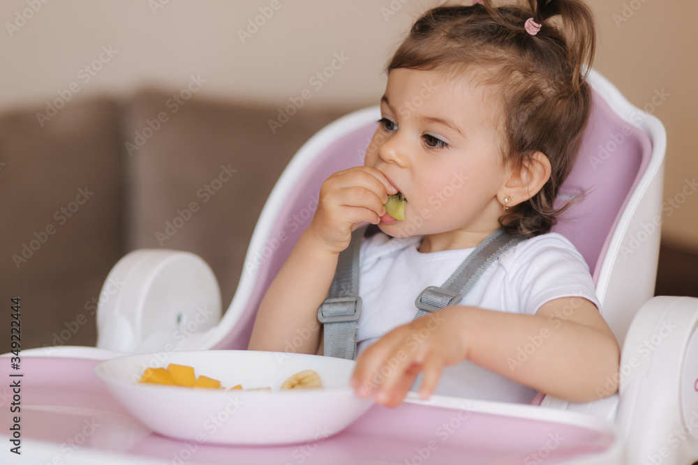 Portrait of happy young baby girl in high chair eating exotic fruits. Healthy nutrition for kids Portrait of happy young baby girl in high chair eating exotic fruits. Healthy nutrition for kids