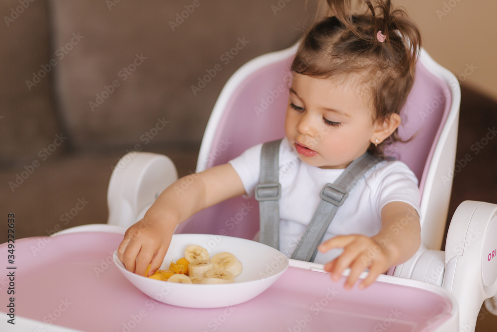 Portrait of happy young baby girl in high chair eating exotic fruits. Healthy nutrition for kids Portrait of happy young baby girl in high chair eating exotic fruits. Healthy nutrition for kids