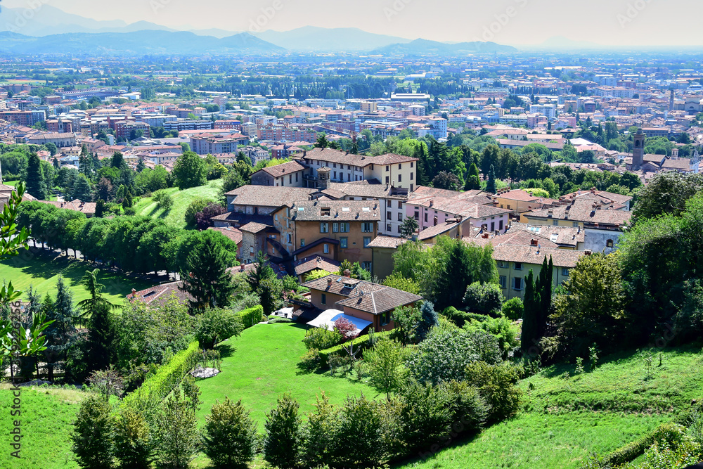 Bergamo / Lombardy, Italy - Panorama of the city of Bergamo, houses with brown tiled roofs, green fields and hills with trees.