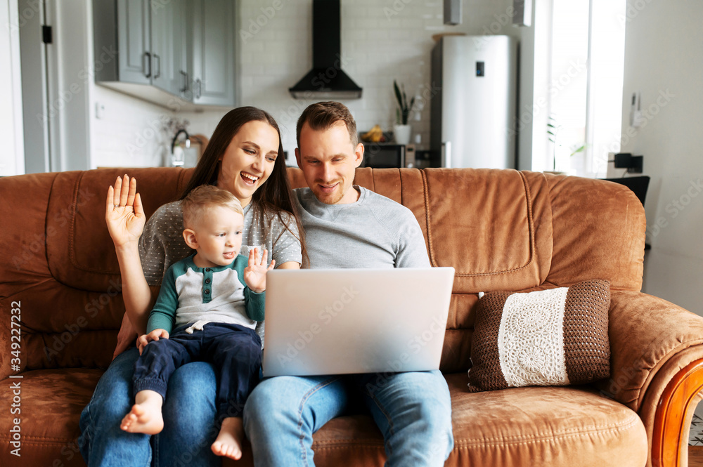 Video call. A young family communicating via video with grandparents. Parents with a baby son sit on couch at home with a laptop and talking online Video call. A young family communicating via video with grandparents. Parents with a baby son sit on couch at home with a laptop and talking online
