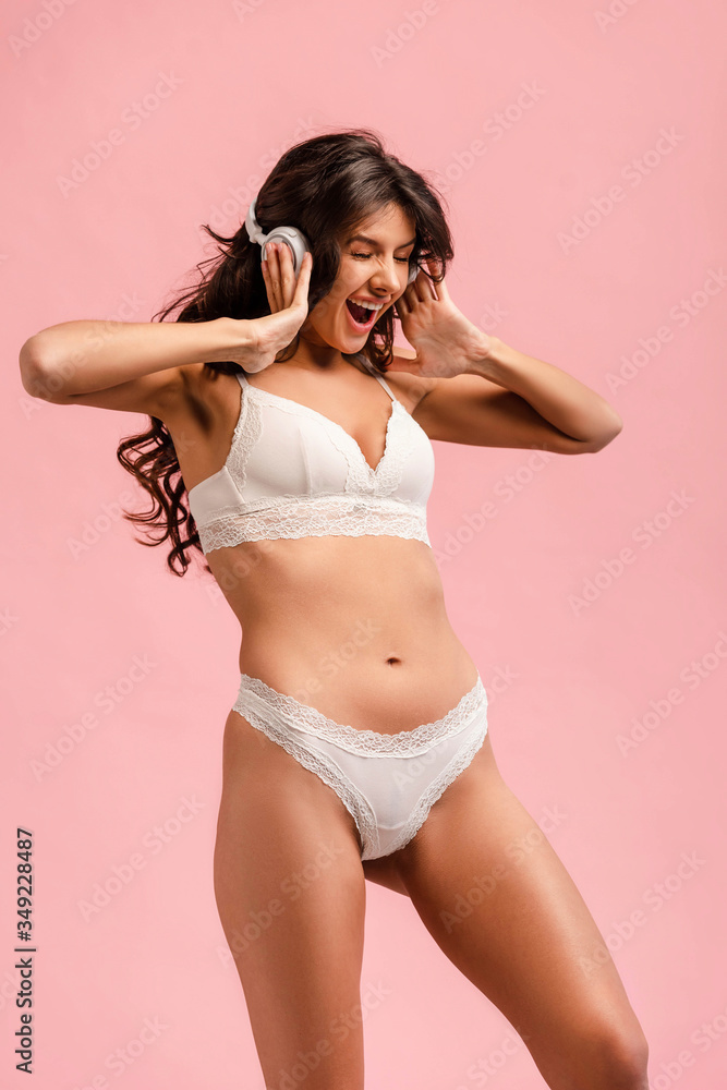 Obraz premium Excited woman in lingerie touching headphones and listening music isolated on pink