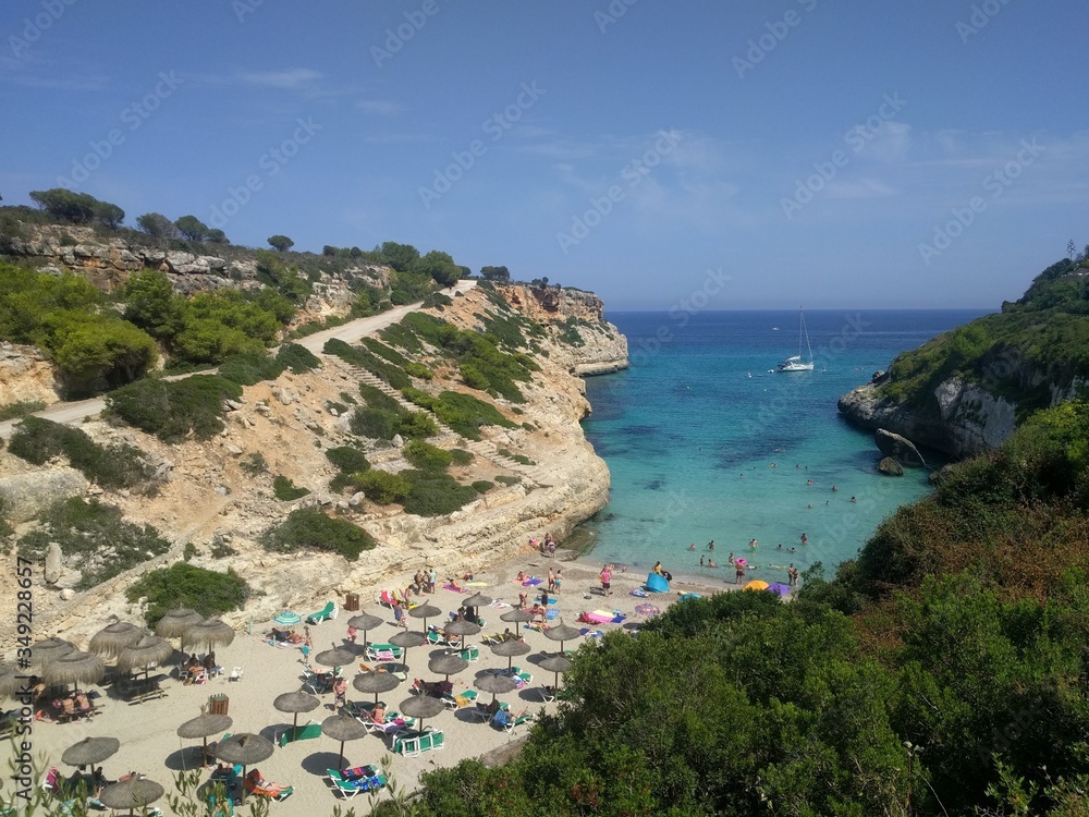 View from the high shore to the sandy beach in the bay of Cala Antena on the island of Mallorca, Spain. It’s a hot summer day, a yacht is visible on the horizon, a bright blue sea, people sunbathe