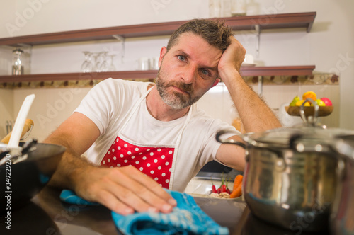 domestic chores stress - young attractive overworked and depressed man in red apron at home kitchen feeling stressed and tired of cooking and dishwashing