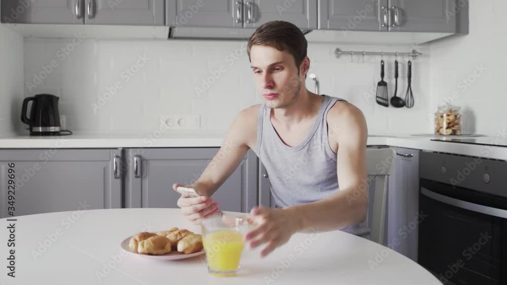 Young man drinking orange juice and using smart device in hand, morning time daily routine of handsome guy sitting at modern kitchen Young man drinking orange juice and using smart device in hand, morning time daily routine of handsome guy sitting at modern kitchen