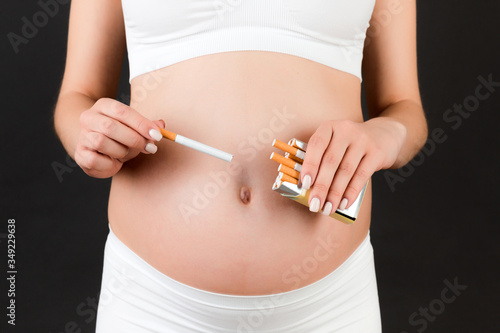 Close of of pregnant woman in white underwear holding a pack of cigarettes at black background. Bad habbit. Toxic influence on a baby
