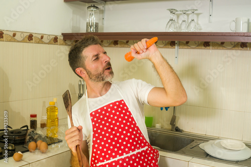 enjoying domestic chores - young cool and attractive man in red apron singing song and dancing crazy happy holding carrot as microphone having fun cooking carefree photo