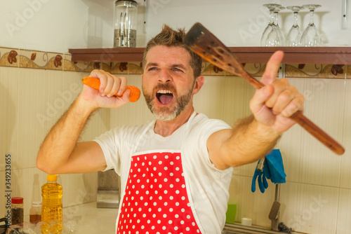 enjoying domestic chores - young cool and attractive man in red apron singing song and dancing crazy happy holding carrot as microphone having fun cooking carefree photo